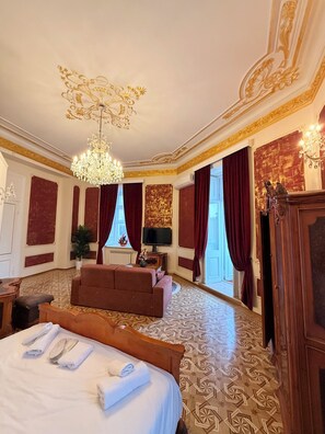 Quarto de hotel com cama, sofá, lustre, televisão e uma janela grande com cortinas.