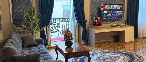 Ein Hotelzimmer mit einer Couch, einem Bett mit blauer Bettdecke, einem runden Teppich, einem kleinen Tisch mit einer Vase und einem Fernseher, der verschiedene Sender zeigt.