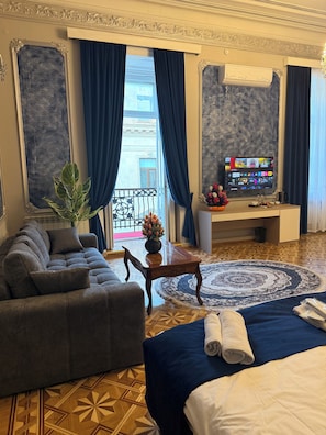 Une chambre d’hôtel avec un canapé, un lit recouvert d’une courtepointe bleue, un tapis rond, une petite table avec un vase et un téléviseur diffusant différentes chaînes.