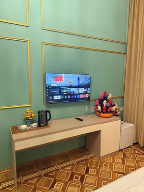 A hotel room with a desk, a television displaying a show, a coffee maker, a vase with flowers, and a bed. - Crown Palace Hotel Baku (Baku)