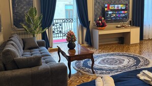 Quarto de hotel com sofá, cama com colcha azul, tapete redondo, pequena mesa com um vaso e TV exibindo diversos canais.