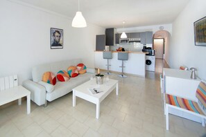Een moderne woonkamer met een bank, salontafel en barstoelen. In de achtergrond is een keuken zichtbaar met een magnetron en wasmachine.