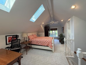 A bedroom with a sloped ceiling, a bed with a checkered bedspread, a desk with a chair, a ceiling fan, and a window. - Bourbon Trail Experience (Louisville)