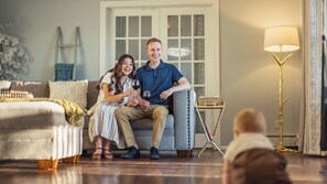A couple sitting on a grey sofa with a small side table, a lamp, and a window in the background.