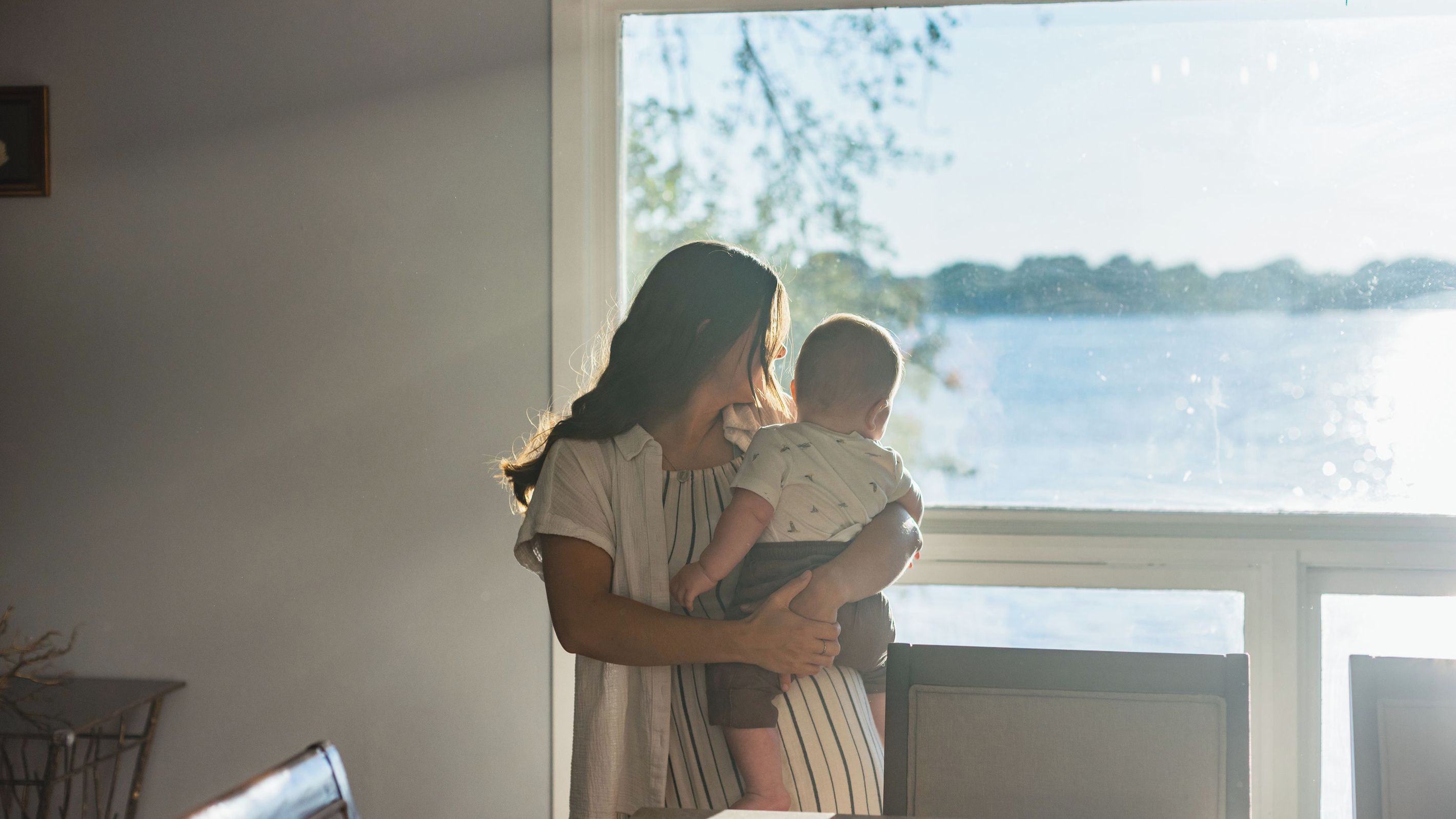 A woman holding a baby by a window with a view of a body of water.