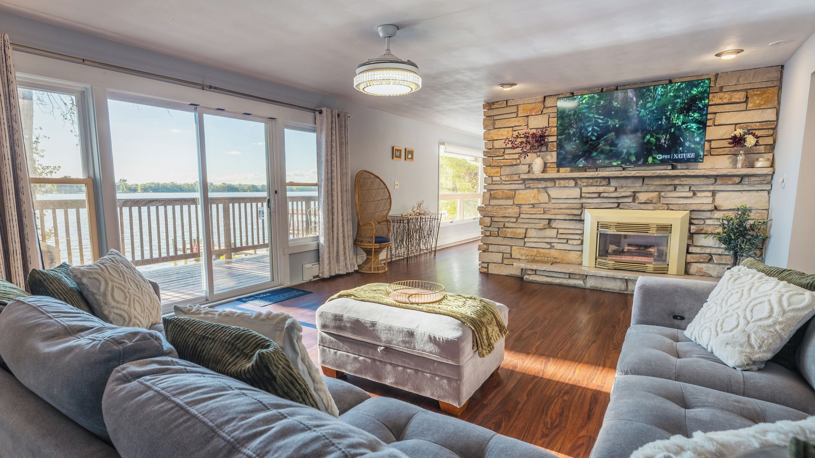A spacious living room with a stone fireplace, a large flat-screen TV mounted on the wall, and a balcony with a view of the water.