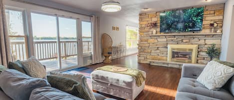 A spacious living room with a stone fireplace, a large flat-screen TV mounted on the wall, and a balcony with a view of the water.