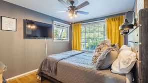 A bedroom with a bed, a ceiling fan, a TV, and a window with yellow curtains.