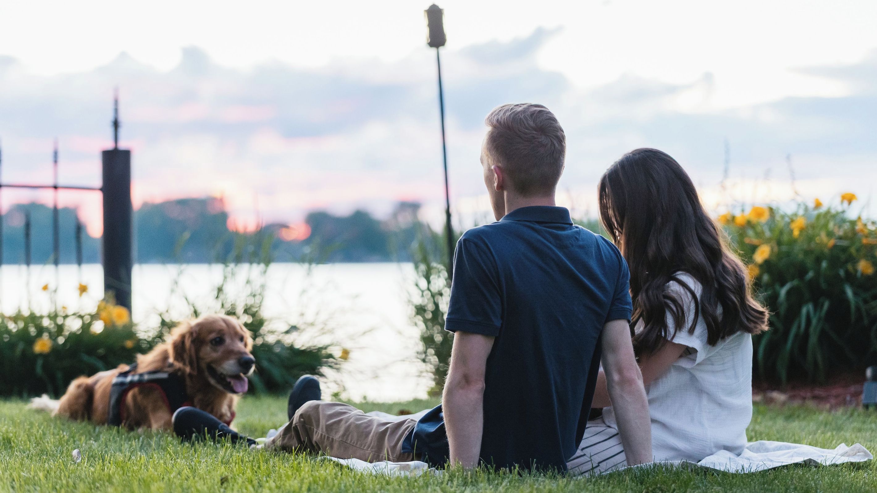 Un couple est assis sur l’herbe avec un chien, et ils ont devant eux une étendue d’eau et un pont.