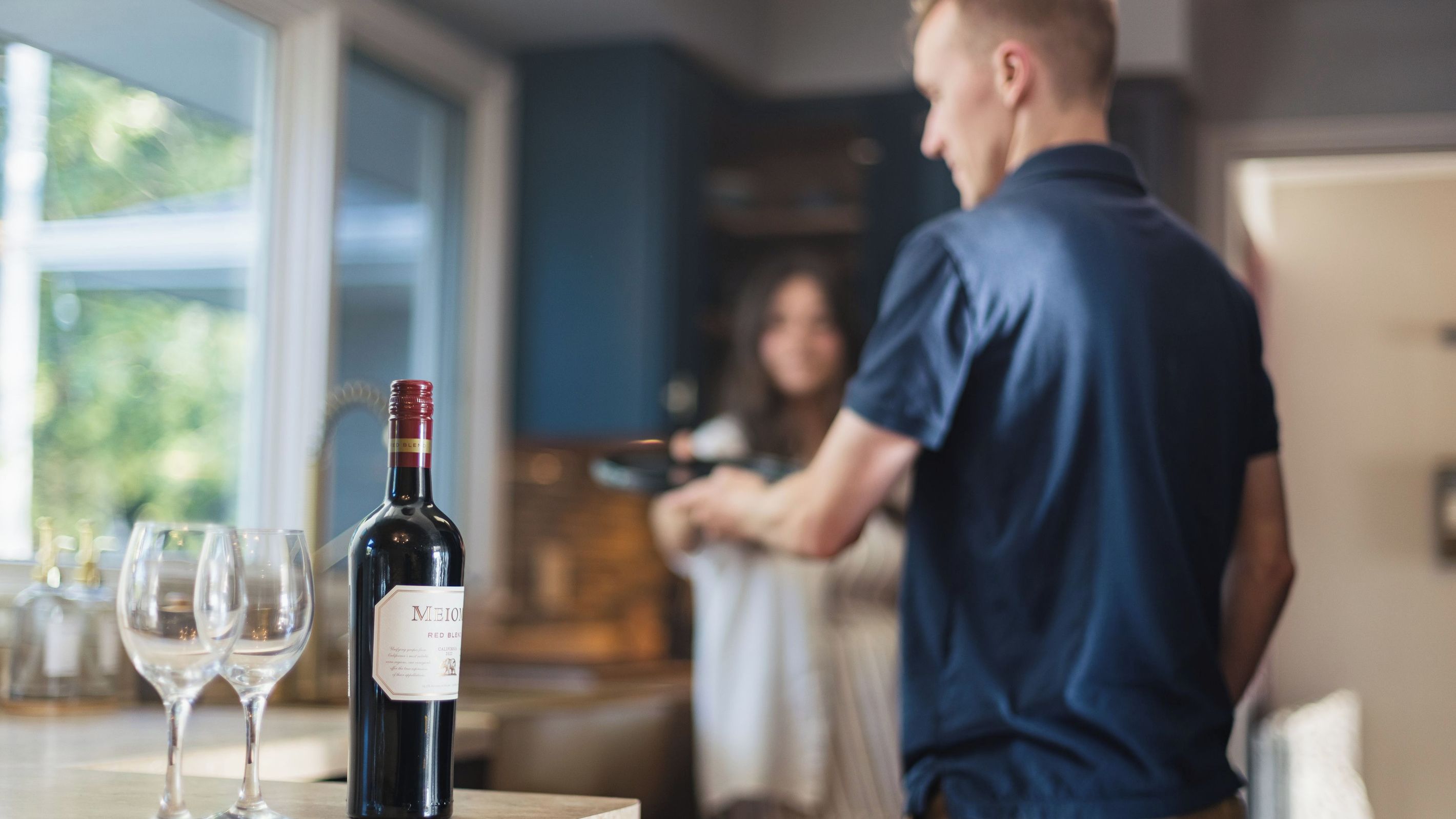 Una botella de vino sobre una mesa con dos copas, un hombre con camisa azul y una mujer al fondo.