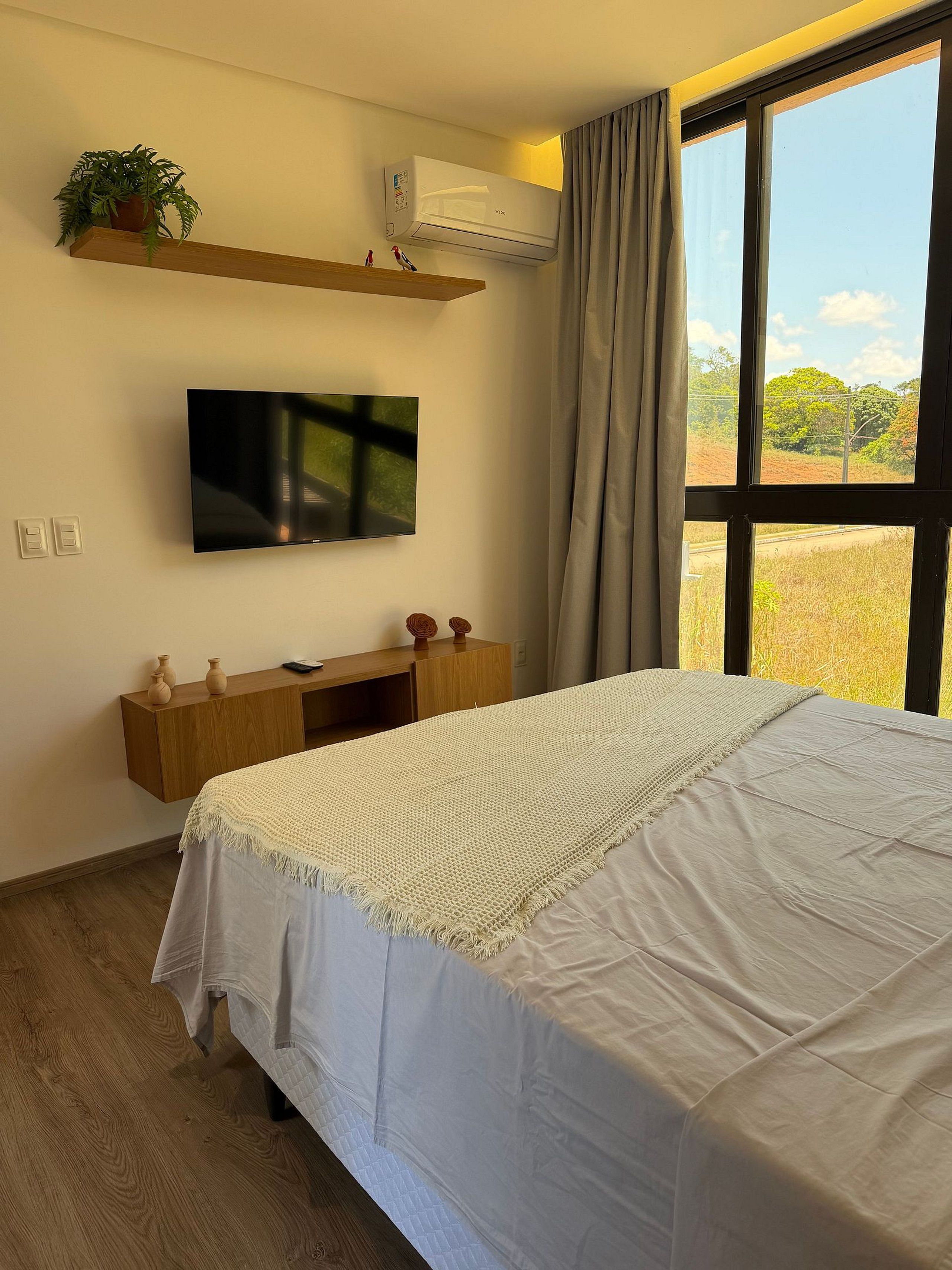 A bedroom with a bed, a TV mounted on the wall, a wooden desk, a potted plant, and a window with curtains.