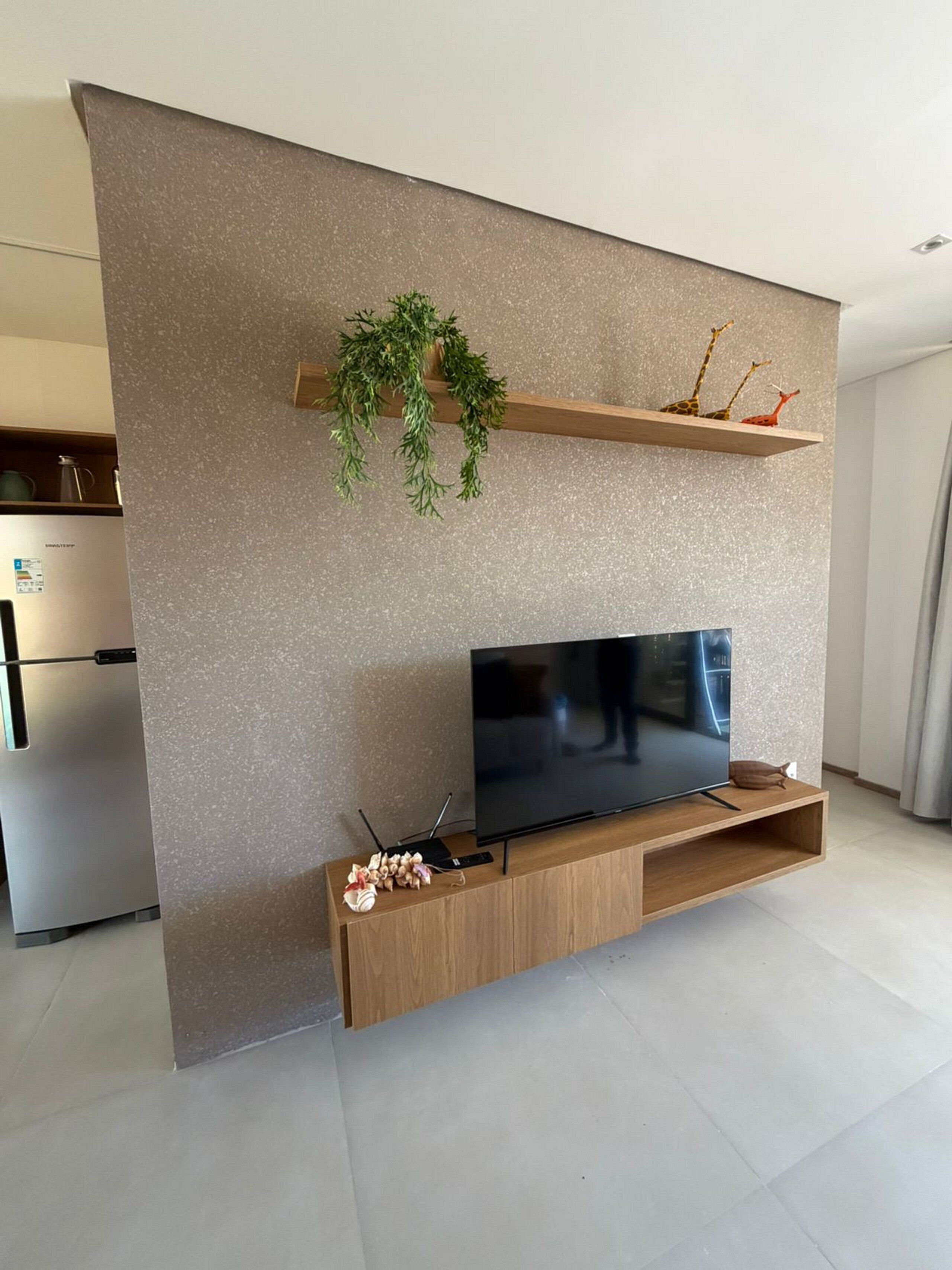 A modern living room with a flat-screen TV on a wooden stand, a small shelf with decorative items, and a refrigerator in the background.