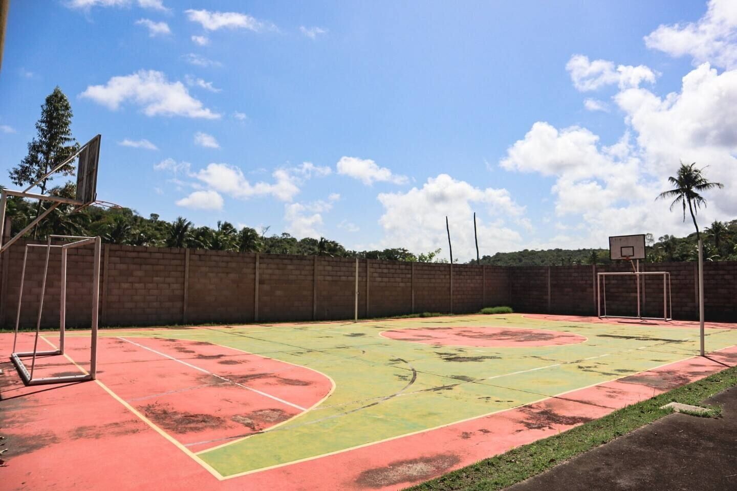 An outdoor basketball court with a red and green playing surface, surrounded by a brick wall and palm trees.