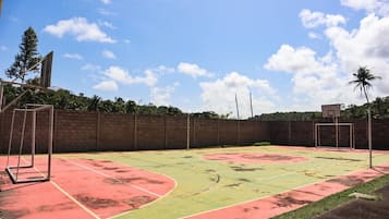 An outdoor basketball court with a red and green playing surface, surrounded by a brick wall and palm trees.
