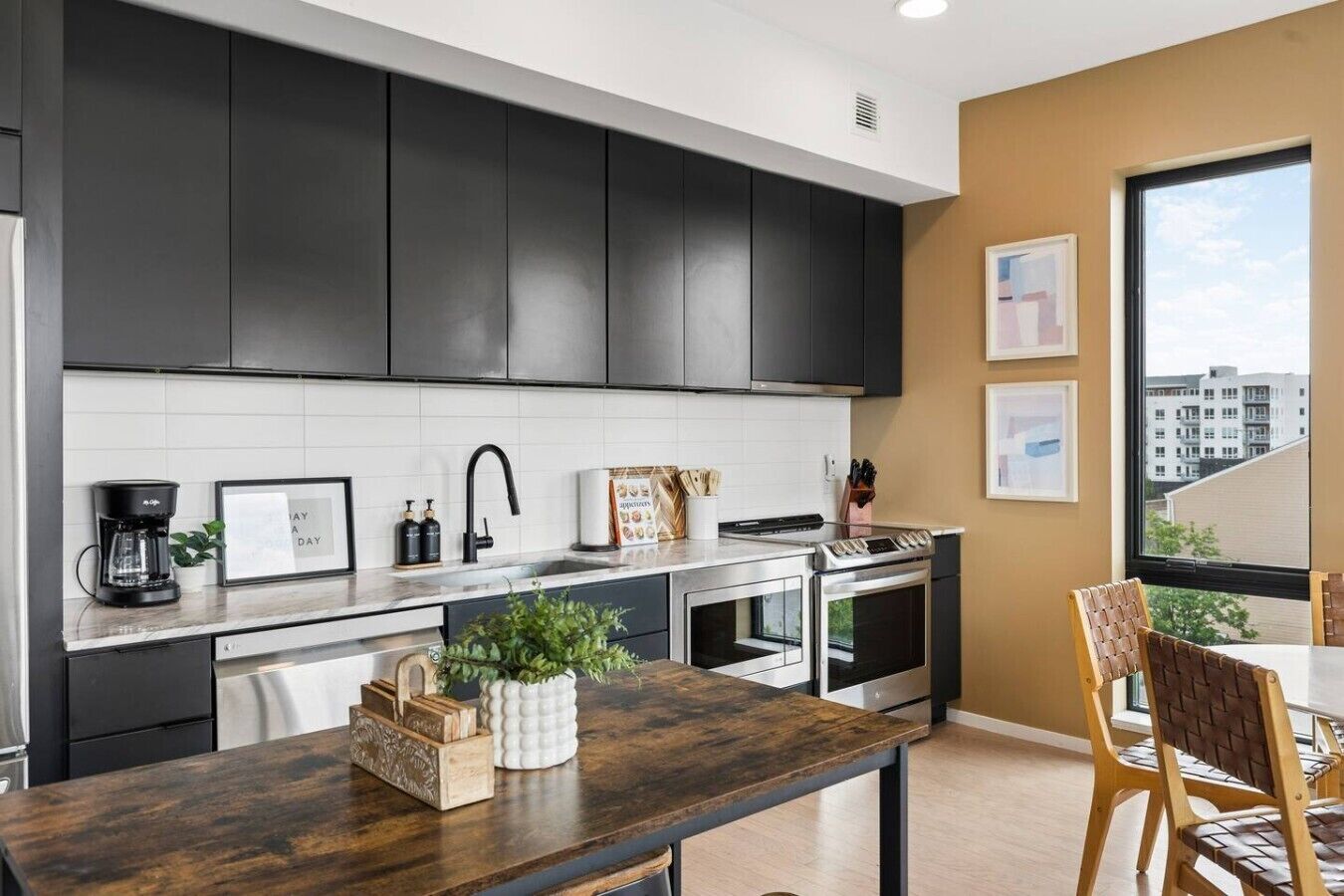 Cocina moderna con gabinetes oscuros, salpicadero blanco y electrodomésticos de acero inoxidable. Hay una zona de comedor con una mesa y sillas de madera.