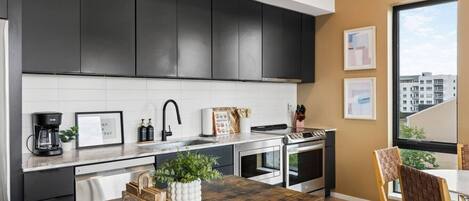 Cocina moderna con gabinetes oscuros, salpicadero blanco y electrodomésticos de acero inoxidable. Hay una zona de comedor con una mesa y sillas de madera.