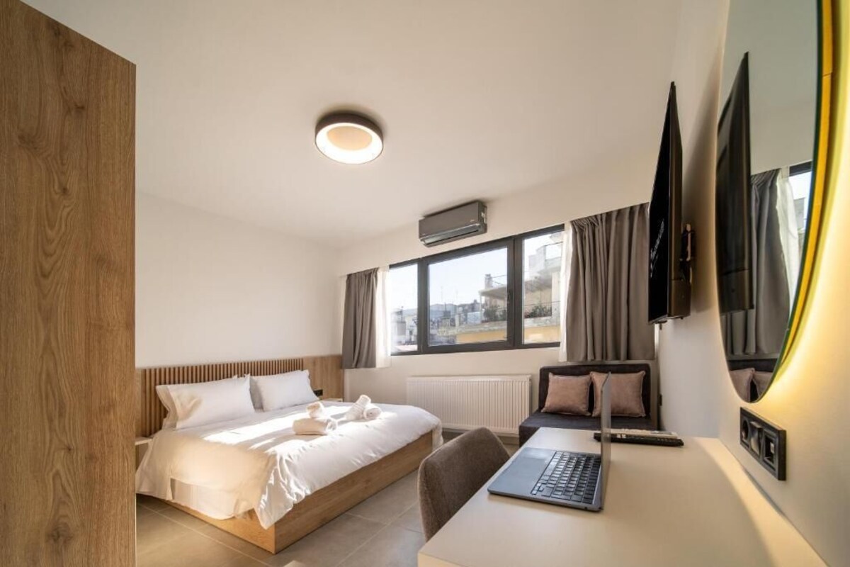 A modern hotel room with a bed, a desk with a laptop, a TV, and a window with curtains.
