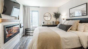 Habitación de hotel con cama, chimenea, televisor, ventana con cortinas, espejo redondo y lámpara de pared.