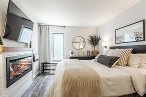 Ein Hotelzimmer mit Bett, Kamin, Fernseher, einem Fenster mit Vorhängen, einem runden Spiegel und einer Wandlampe.