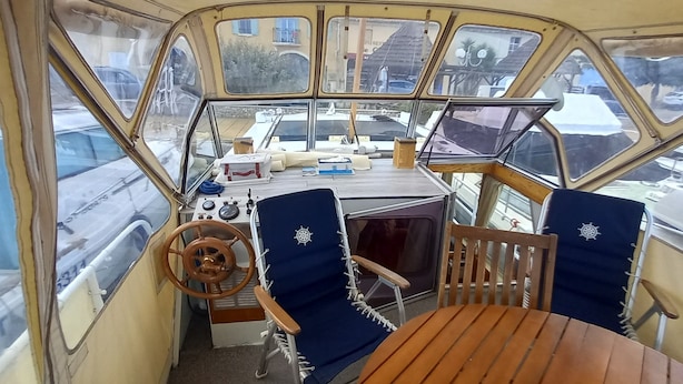 Het interieur van een boot, met een stuurwiel, twee blauwe stoelen, een tafel en een klein cabinegedeelte.