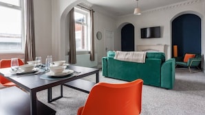 Una habitación con una mesa de comedor dispuesta para cuatro personas, un sofá verde, sillas naranjas, un televisor y una chimenea.
