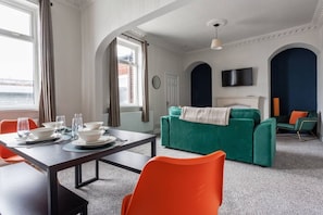 Ein Zimmer mit Esstisch für vier Personen, einem grünen Sofa, orangefarbenen Sesseln, einem Fernseher und einem Kamin.
