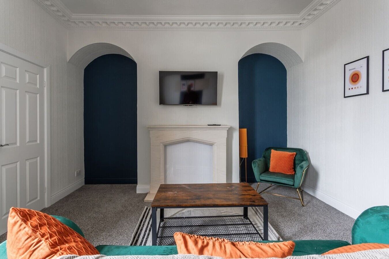 Ein Wohnzimmer mit Kamin, einem darüber hängenden Fernseher und einem grünen Sessel mit orangefarbenem Kissen.