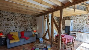 A rustic interior with exposed wooden beams, a stone wall, a sofa with colorful cushions, a small round table, and a kitchen area with a checkered tablecloth. - “L'air De Vacances” Cottage with Private Terrace and Wi-Fi (Bonnée)