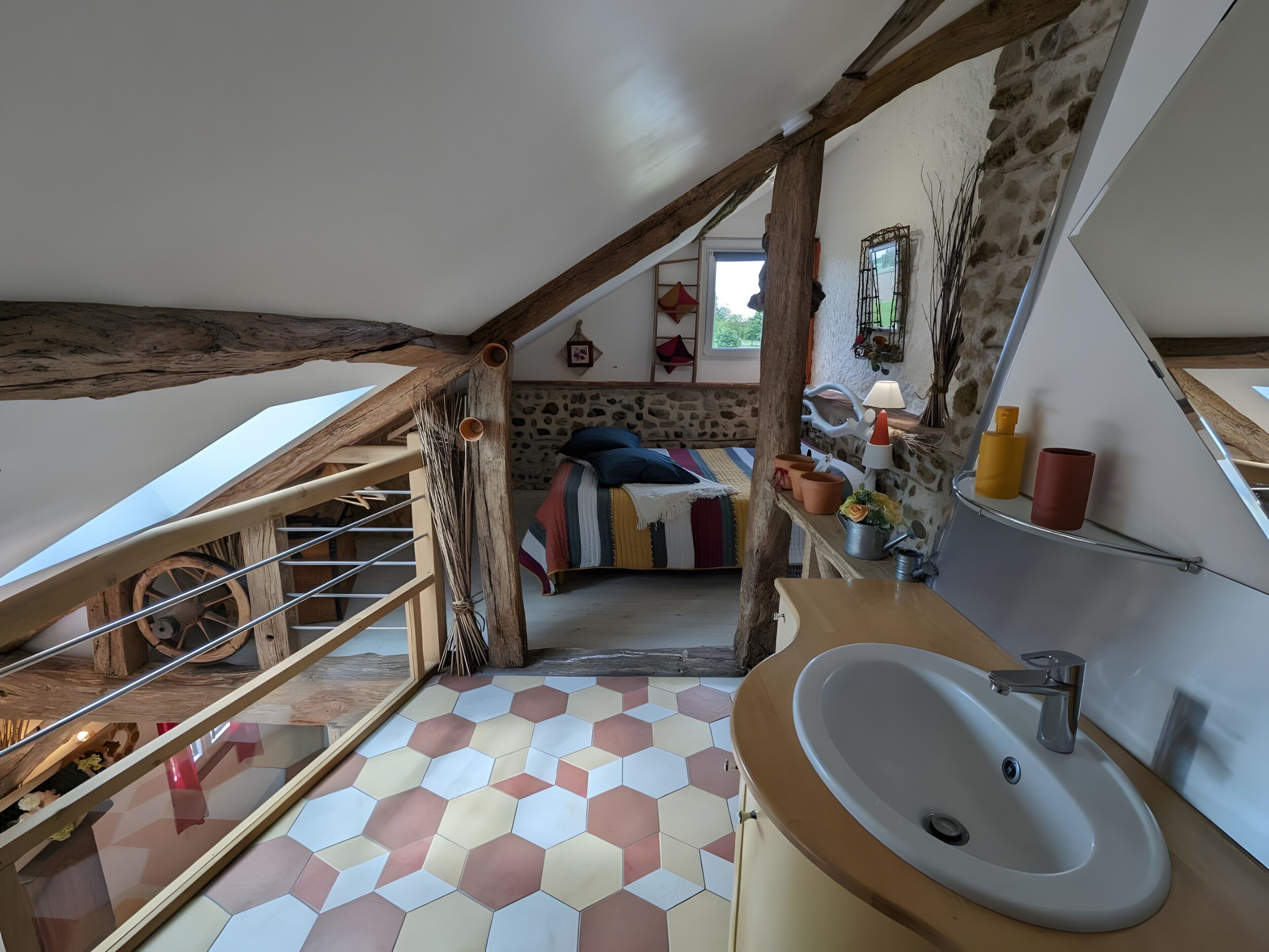 Une chambre avec un plafond en pente, un escalier en bois, un lit avec une literie colorée, un mur en pierre et une salle de bain avec un lavabo et un miroir.
