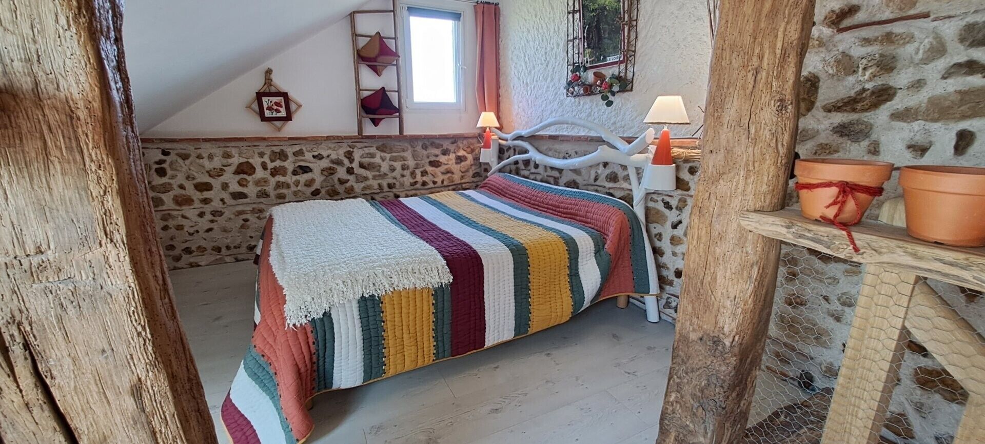 Un dormitorio acogedor con pared de piedra, una cama con manta a rayas de colores, cabecera de madera y una mesita con plantas en macetas.