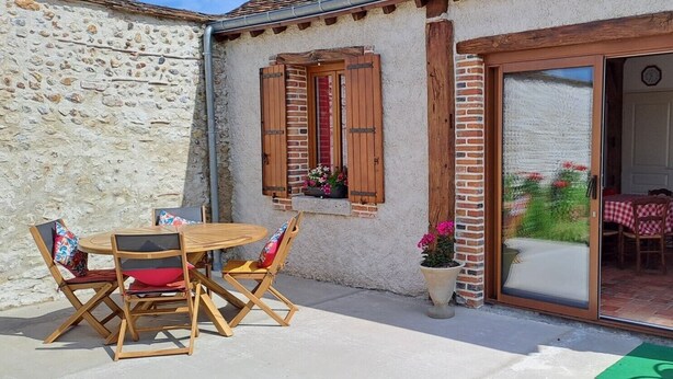 A stone building with a wooden table and chairs, a sliding glass door leading to a kitchen, and a small balcony with a potted plant. - “L'air De Vacances” Cottage with Private Terrace and Wi-Fi (Bonnée)