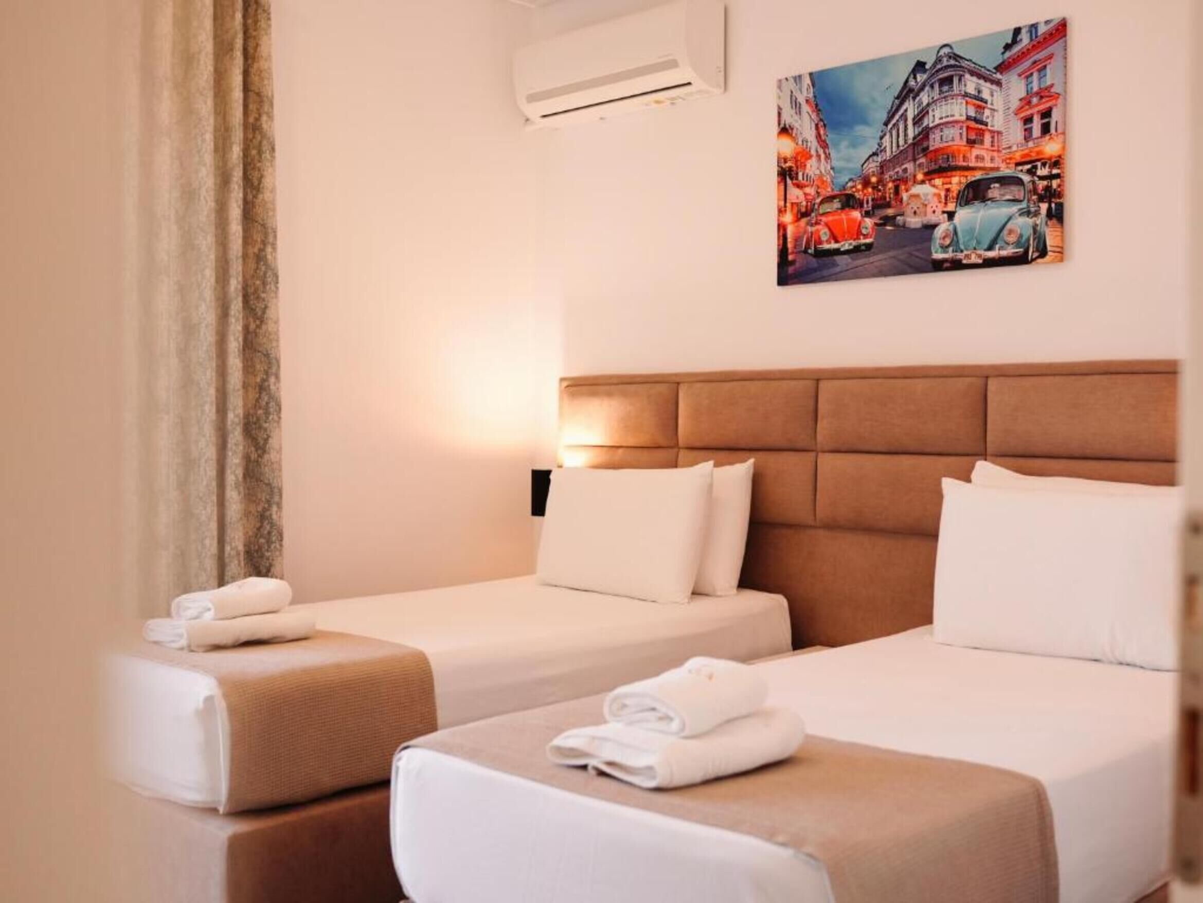 Une chambre d’hôtel avec deux lits, une tête de lit, un appareil de climatisation et un tableau représentant une scène de rue.