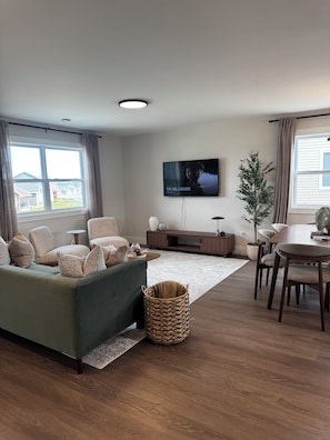 Ein modernes Wohnzimmer mit einer Couch, Fernseher, Esstisch und einem großen Fenster.