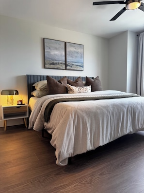A bedroom with a bed, nightstand, lamp, ceiling fan, and two wall-mounted paintings.