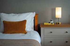Uma cama com roupa de cama branca e um travesseiro marrom, uma mesa de cabeceira com abajur, e uma pequena estatueta.