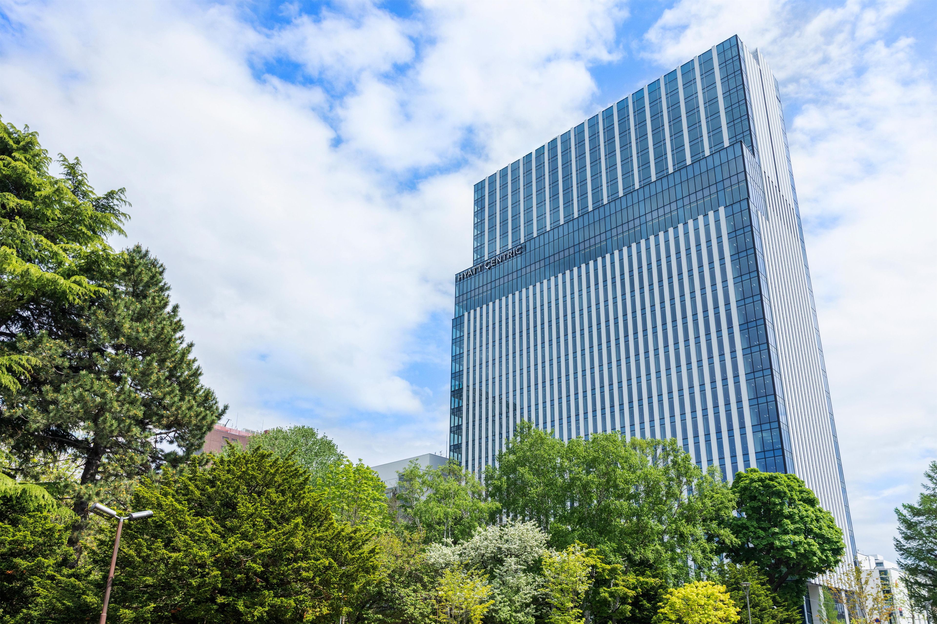 A modern high-rise building with a glass facade, surrounded by lush green trees.