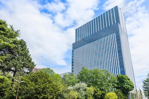 A modern high-rise building with a glass facade, surrounded by lush green trees.