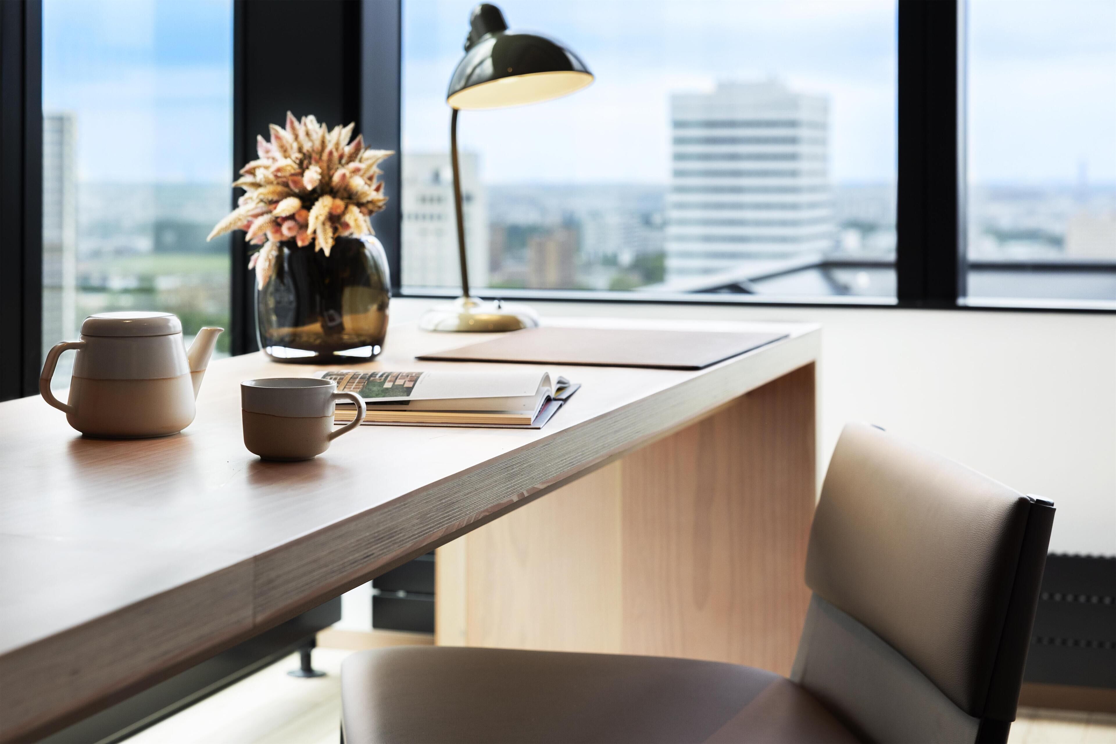 A modern office desk with a laptop, a vase with dried flowers, a lamp, and a view of the city through large windows.