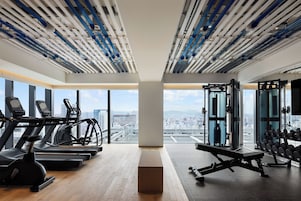 A gym with treadmills, a weight bench, and a squat rack, featuring large windows and a city view.
