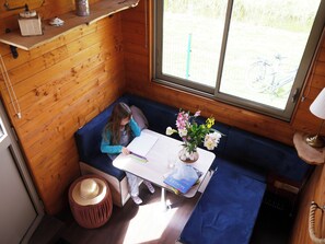 L’intérieur d’un chalet en bois, avec un banc bleu rembourré, une table ornée d’un vase de fleurs et une fenêtre donnant sur un paysage verdoyant.