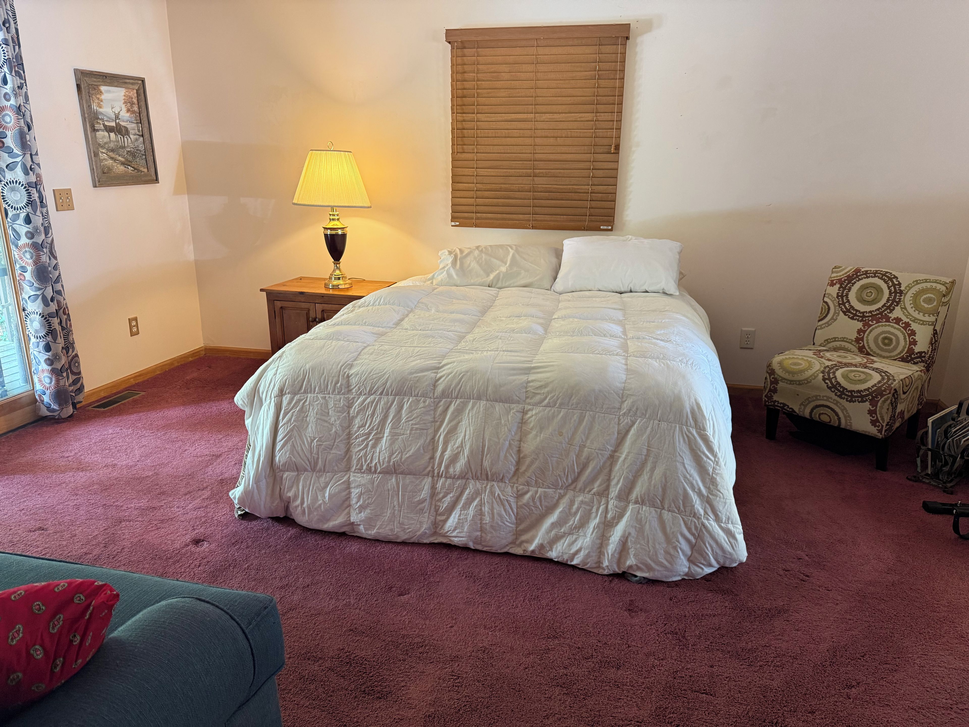A bedroom with a large bed, a bedside lamp, a chair, and a window with blinds.