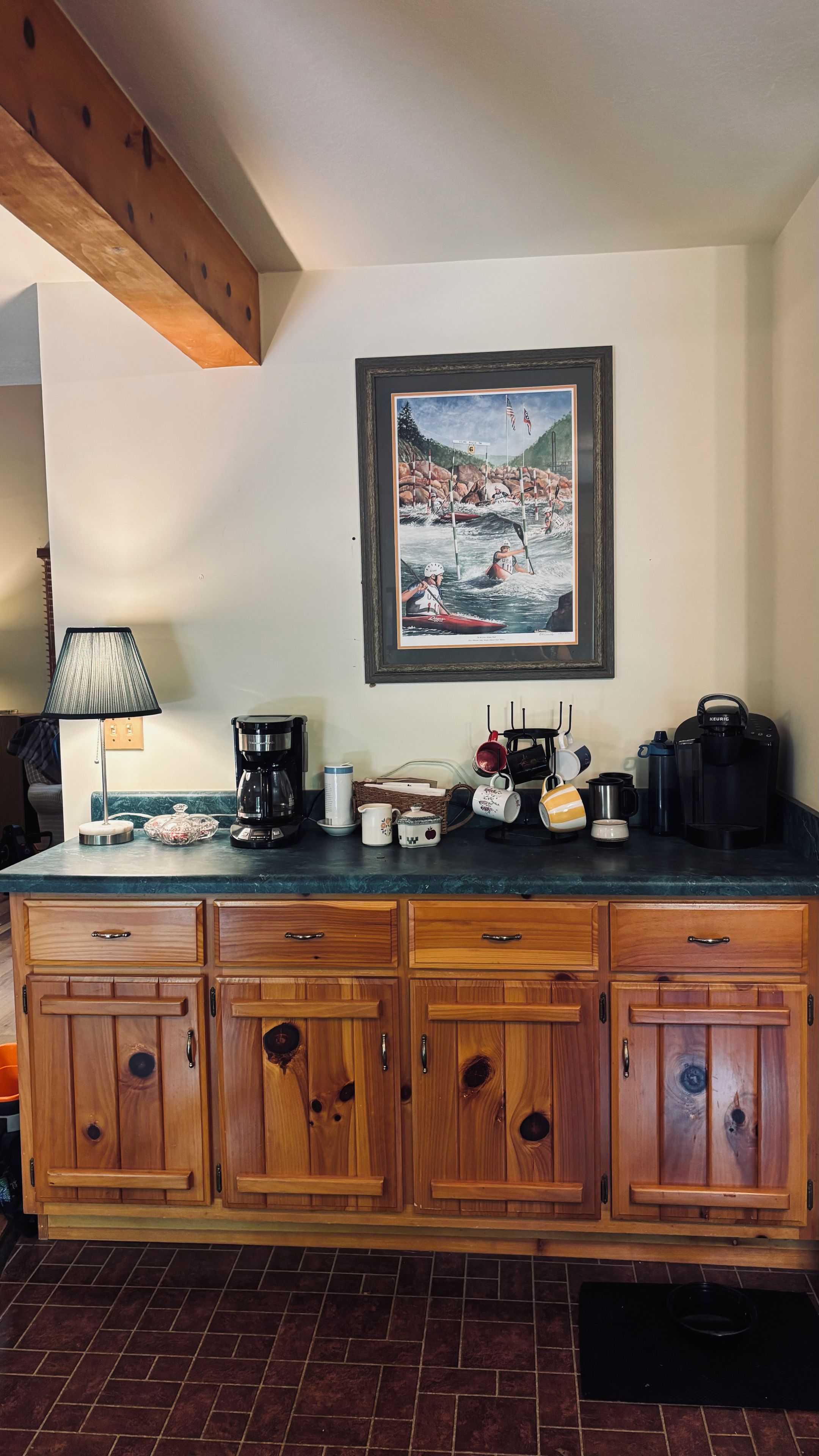 Une cuisine avec des meubles de rangement en bois, une machine à café, des tasses et un tableau encadré au mur.
