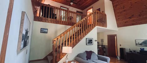 A spacious living area with a wooden staircase, a grey sofa, a small table with a lamp, and a television.