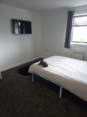 Une chambre à coucher comprenant un lit, un téléviseur à écran plat fixé au mur, une fenêtre avec des rideaux et un radiateur.
