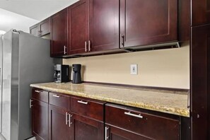 A kitchen with dark wooden cabinets, a granite countertop, a refrigerator, and a coffee maker. - Villa Varges - Be our Guest! (Lehigh Acres)