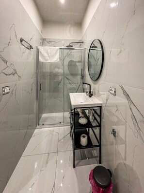 Une salle de bain moderne avec une douche à parois de verre, un lavabo blanc avec un robinet noir, un miroir et une étagère noire avec un porte-rouleau de papier toilette.