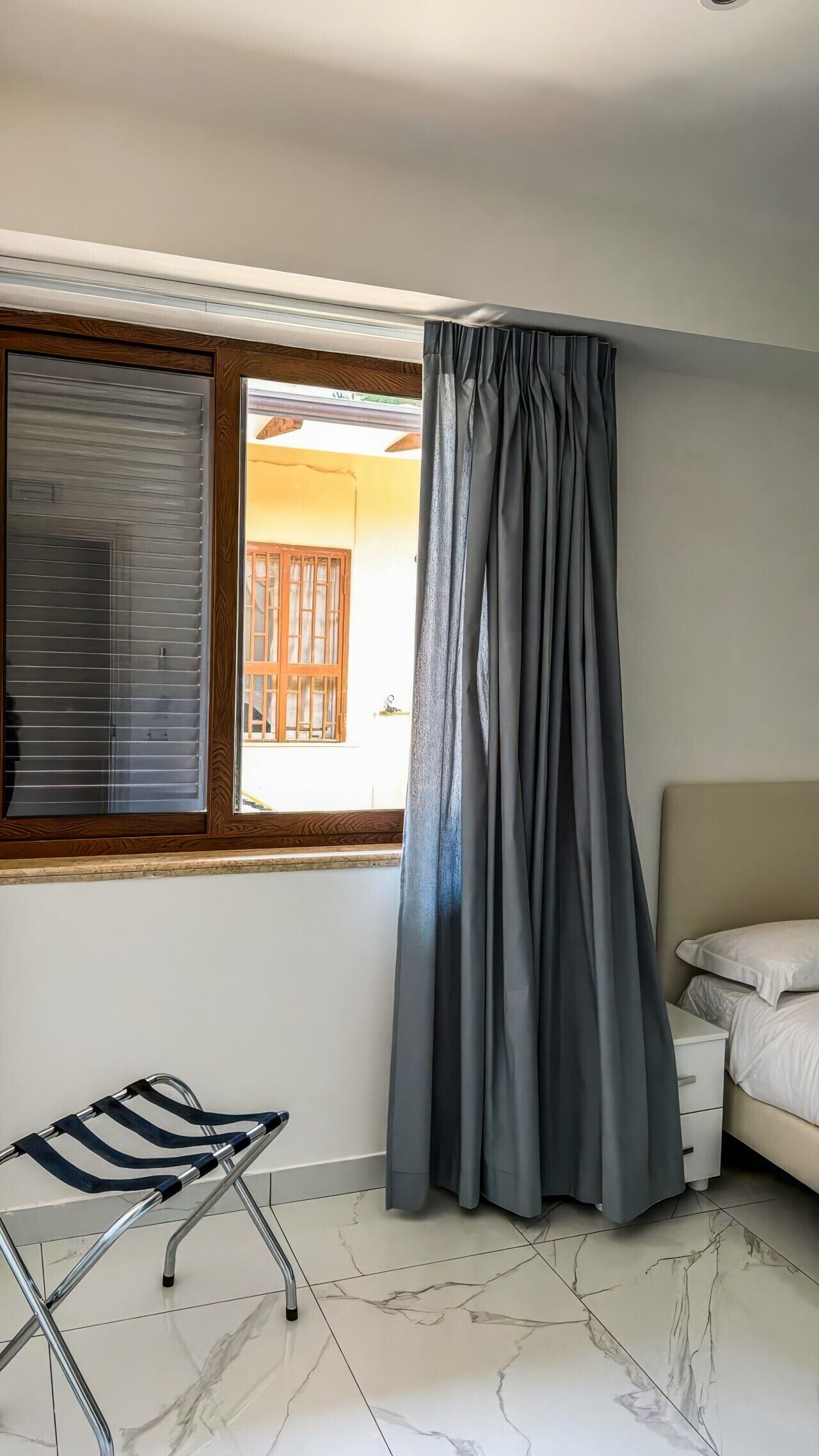 Una camera da letto con un letto, una finestra con tende, una tenda, una sedia e un pavimento piastrellato.