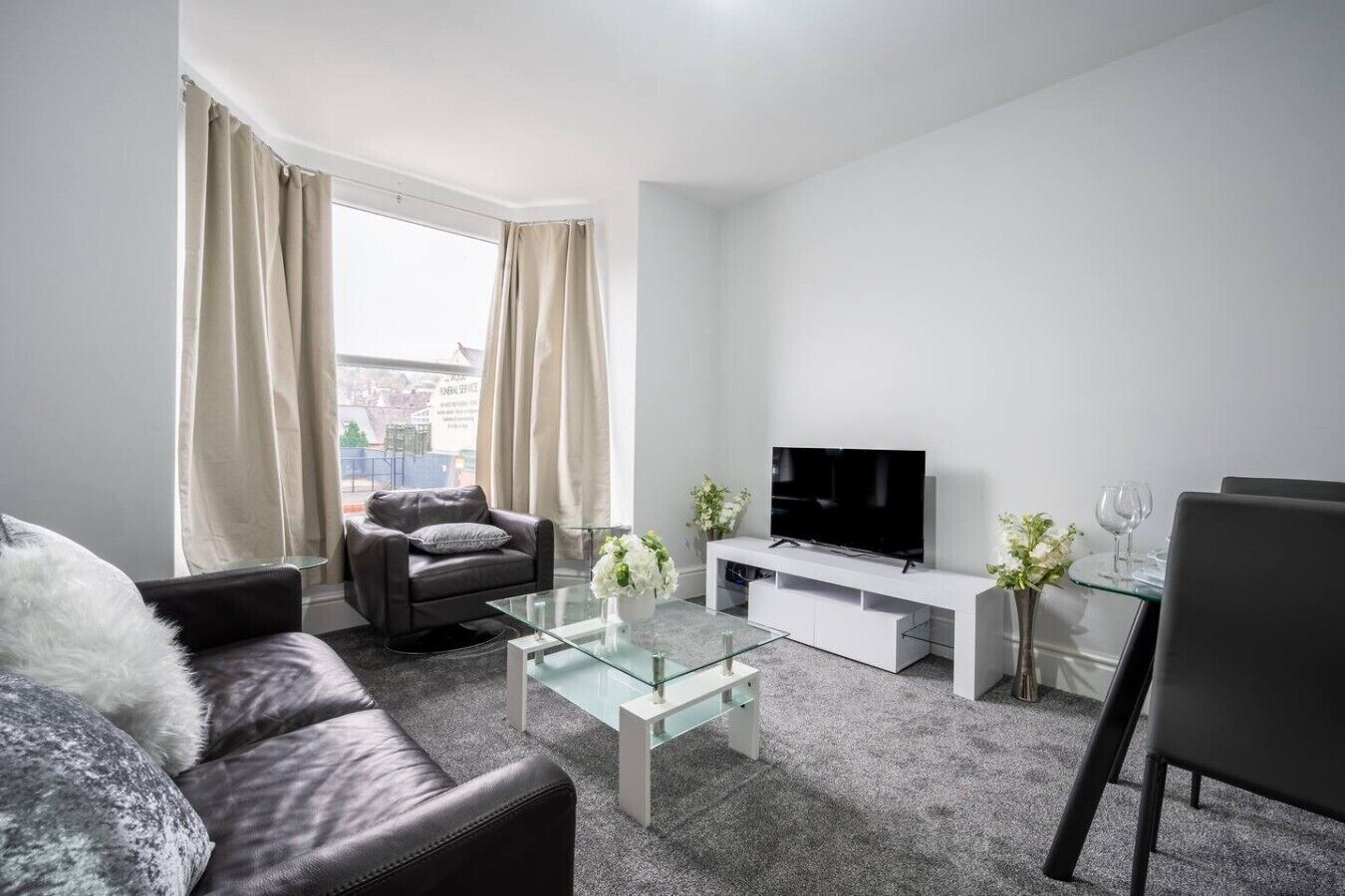 A modern living room with a sofa, a glass coffee table, a TV on a white stand, and a dining area with chairs and a glass table.