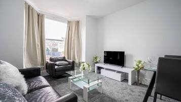 A modern living room with a sofa, a glass coffee table, a TV on a white stand, and a dining area with chairs and a glass table.