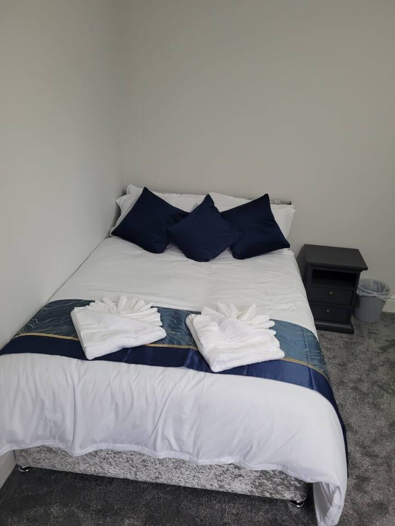 Un lit bien fait, avec une couette blanche et bleue, trois oreillers bleu foncé et deux serviettes blanches pliées.